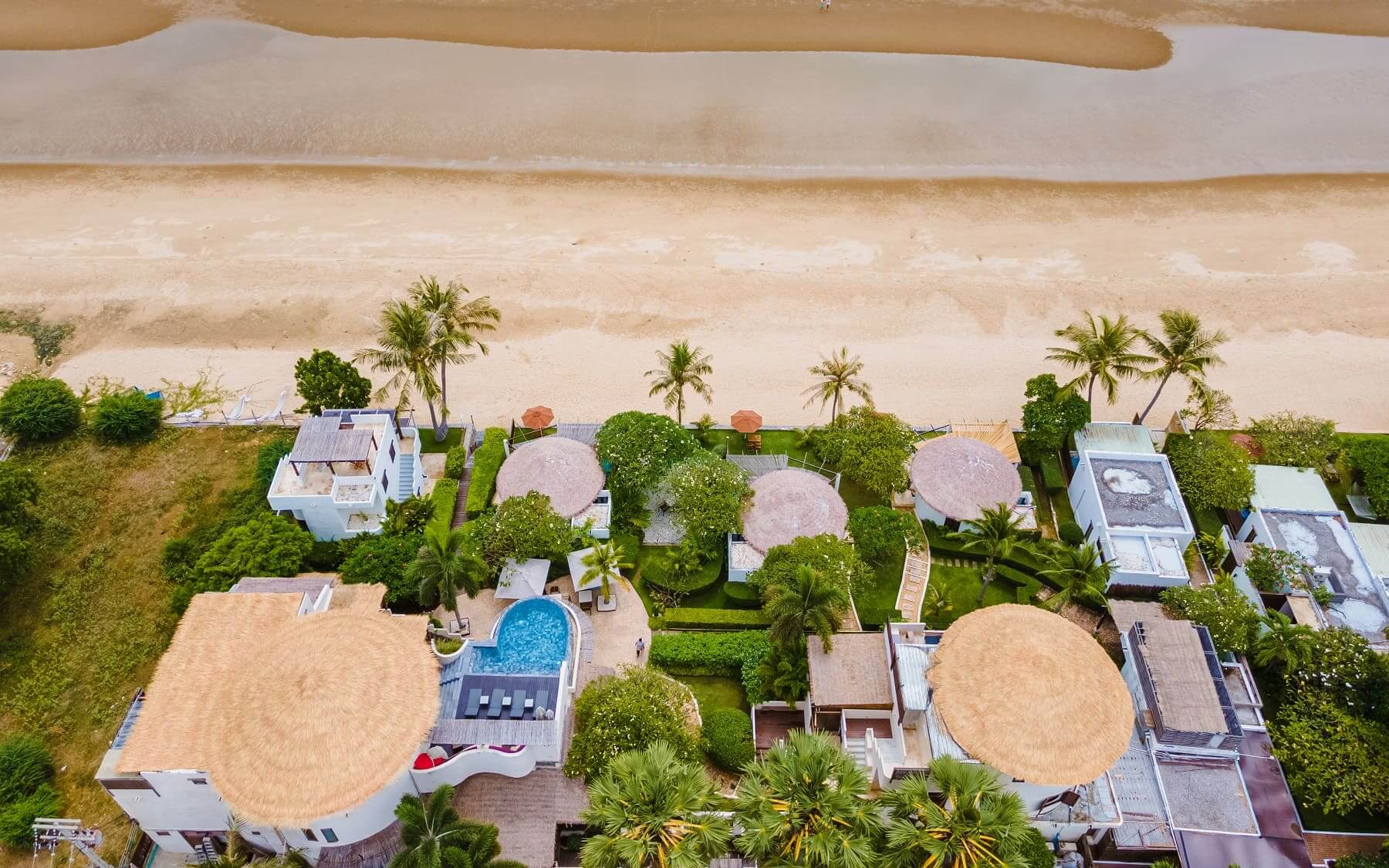 Travel X Aerial view of beachfront resort villas with thatched roofs, swimming pools, palm trees, and a sandy beach adjacent to the ocean. Book your trip now with Travel X for an unforgettable experience. Book Your Trip With Travel X