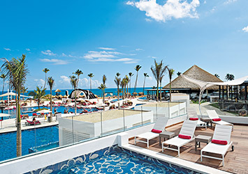 Travel X Resort pool area with lounge chairs and umbrellas, surrounded by palm trees. In the background, guests enjoy the beach and ocean under clear blue skies—perfect for a relaxing vacation with Travel X. Book Your Trip With Travel X