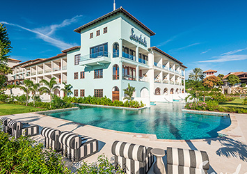 Travel X Luxurious resort with a large pool, surrounded by greenery and featuring a multi-story building with balconies and a sign that reads "Sandals." Perfect for your next Travel X vacation, the sky is clear and blue. Book Your Trip With Travel X