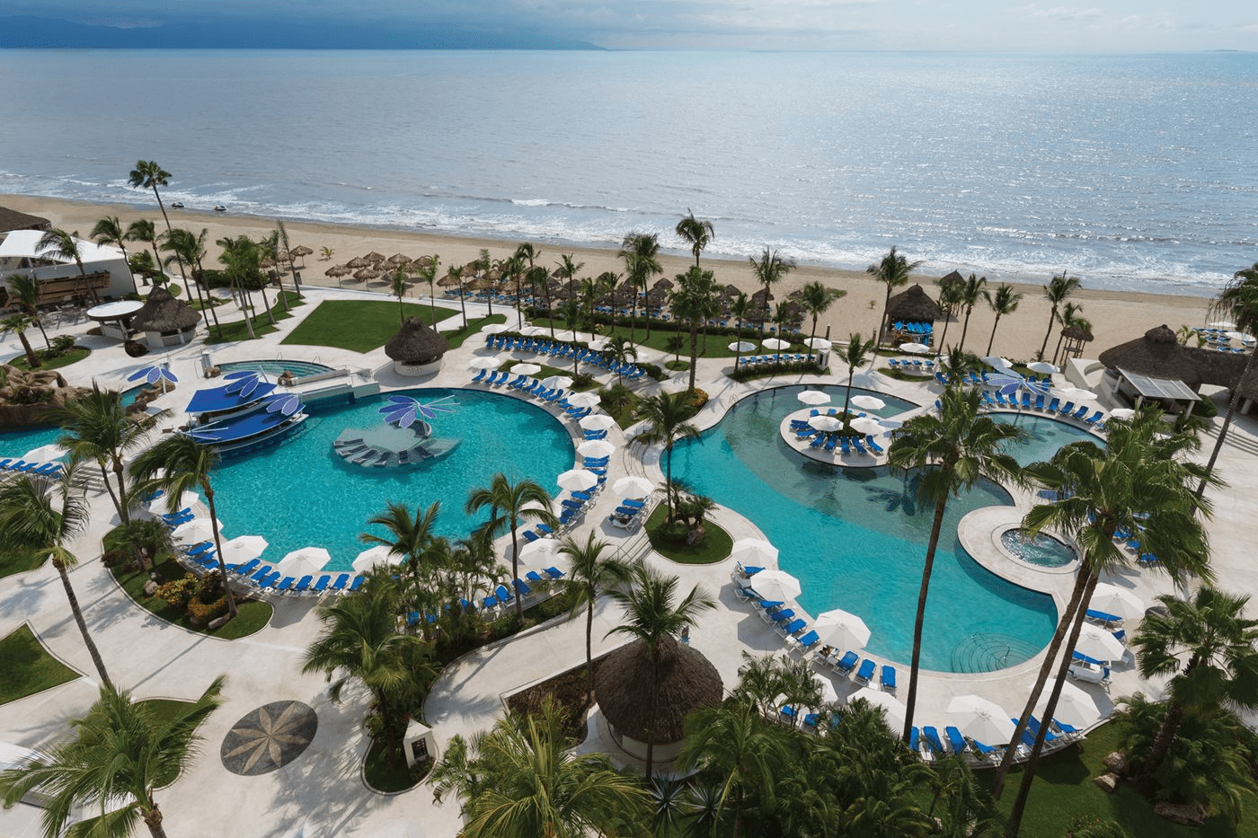 Travel X Aerial view of a beachfront resort featuring multiple swimming pools, sun loungers, palm trees, cabanas, and sandy beach with ocean waves in the background. Perfect for your next vacation; book your trip with Travel X today! Book Your Trip With Travel X