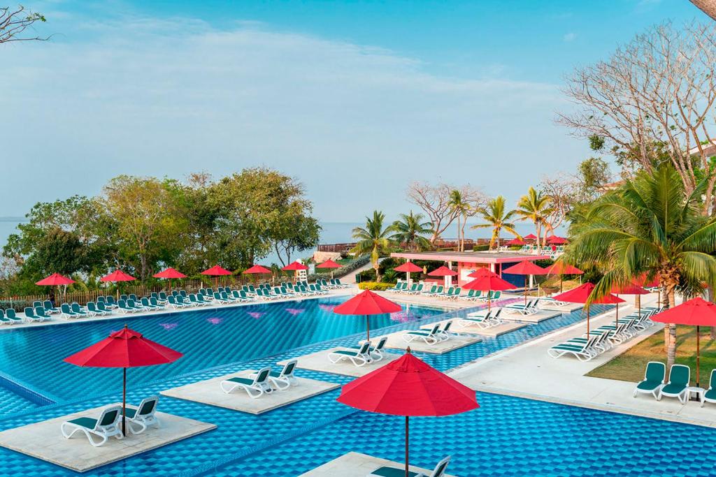 Travel X Outdoor swimming pool with blue tiles, surrounded by lounge chairs and red umbrellas, situated amid tropical trees and clear skies. Book your trip now to this stunning resort for the ultimate vacation experience. Book Your Trip With Travel X