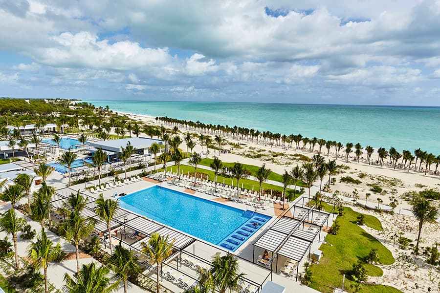 Travel X Aerial view of a beachside resort with multiple pools, rows of lounge chairs, cabanas, palm trees, and a sandy beach leading to a turquoise ocean under a partly cloudy sky. Perfect for your next vacation, Travel X helps you book your trip effortlessly. Book Your Trip With Travel X