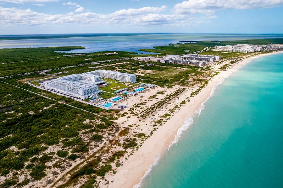 Travel X Aerial view of a coastal resort featuring several white buildings, swimming pools, and a sandy beach adjacent to turquoise waters, with green vegetation in the background. Book your trip now and experience paradise with Travel X! Book Your Trip With Travel X