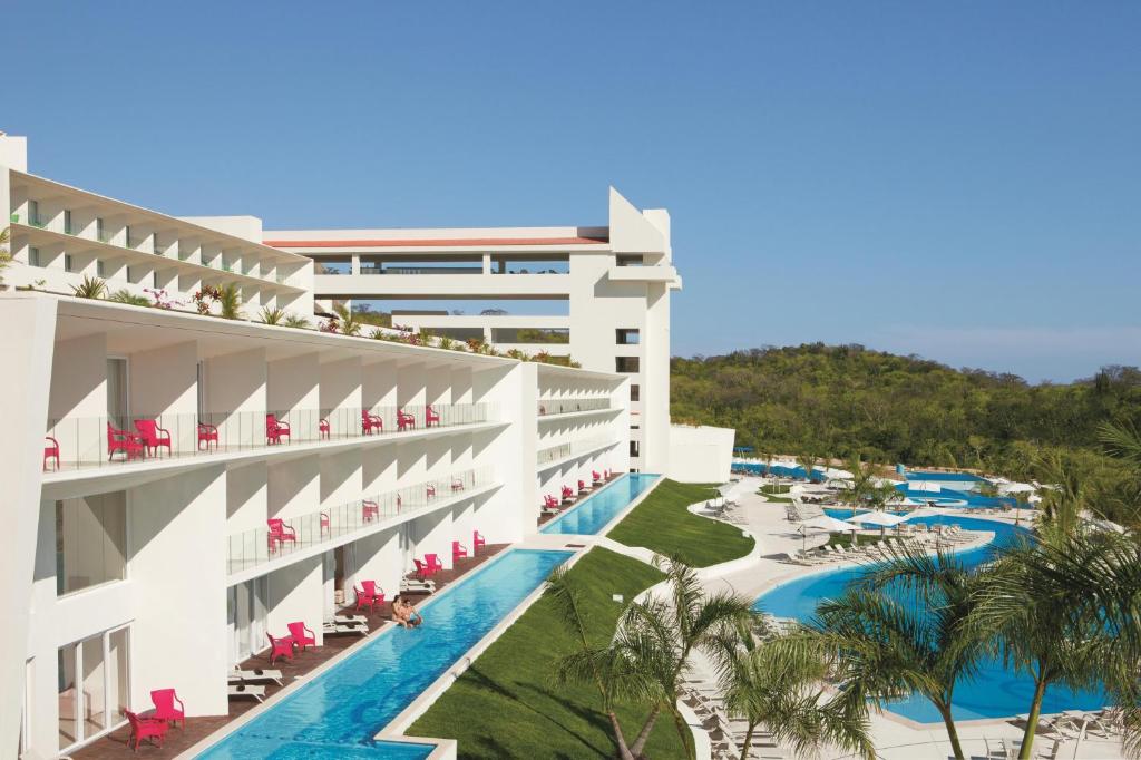 Travel X Modern white building with red chairs on balconies beside a long, narrow pool. The structure overlooks a larger curved pool with white lounge chairs and palm trees, perfect for a Travel X vacation. Blue sky and greenery in the background enhance this stunning resort scene. Book Your Trip With Travel X