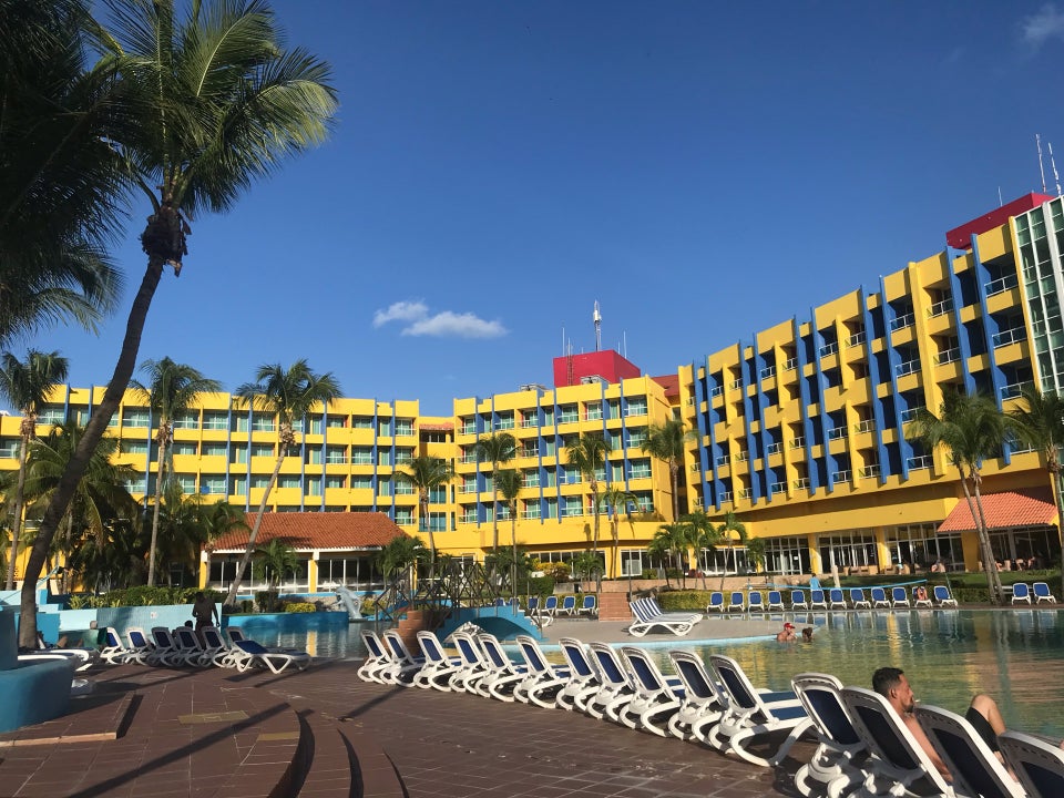 Travel X Book Your Trip to a bright yellow hotel with balconies, surrounded by palm trees. Lounge chairs line the edge of a large pool. Clear blue sky and a few wisps of white clouds are visible above, perfect for your next Vacation with Travel X. Book Your Trip With Travel X