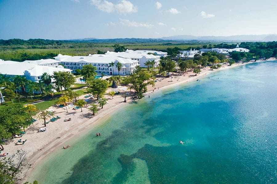 Travel X Aerial view of a coastal resort with white buildings, surrounded by trees, adjacent to a beach with clear blue water and scattered beachgoers. Book your trip now for an unforgettable vacation! Book Your Trip With Travel X