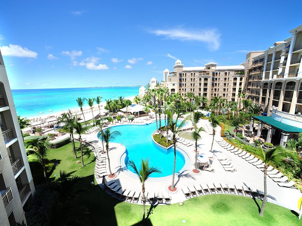 Travel X Aerial view of Travel X's seaside resort with a large pool, palm trees, lounge chairs, and multiple buildings against a backdrop of turquoise ocean and clear blue sky—perfect for your next vacation. Book Your Trip With Travel X