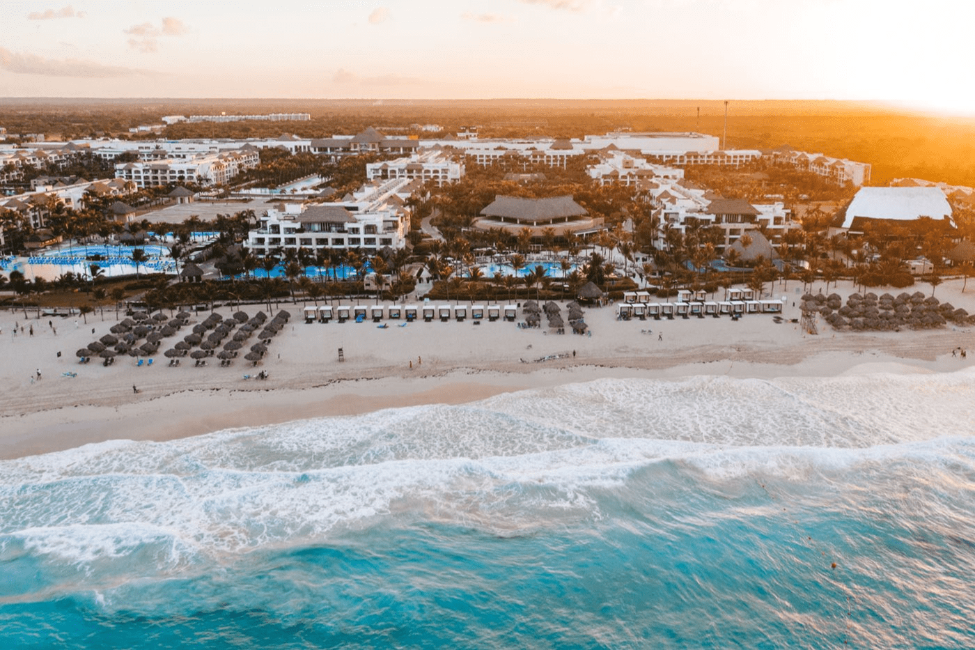 Travel X Aerial view of a beachfront resort with multiple buildings, inviting pools, and shaded lounging areas. The ocean waves gently meet the sandy shore as the sun sets in the background. Book your trip now for an unforgettable vacation experience! Book Your Trip With Travel X