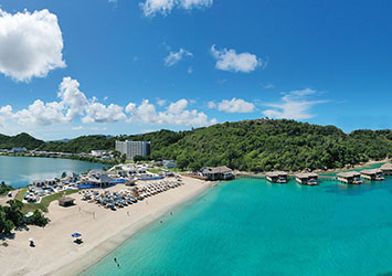 Travel X Aerial view of a beachfront resort with white buildings, sun umbrellas, lush green hills, a clear blue sky, and turquoise water. Book your trip now and dive into the ultimate vacation experience! Book Your Trip With Travel X