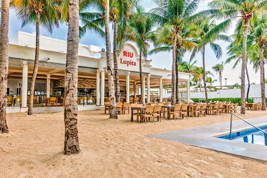 Travel X Outdoor seating area at the RIU Lupita resort surrounded by palm trees, with tables and chairs on sandy ground, and a building in the background. Embark on your dream vacation and book your trip with Travel X today! Book Your Trip With Travel X