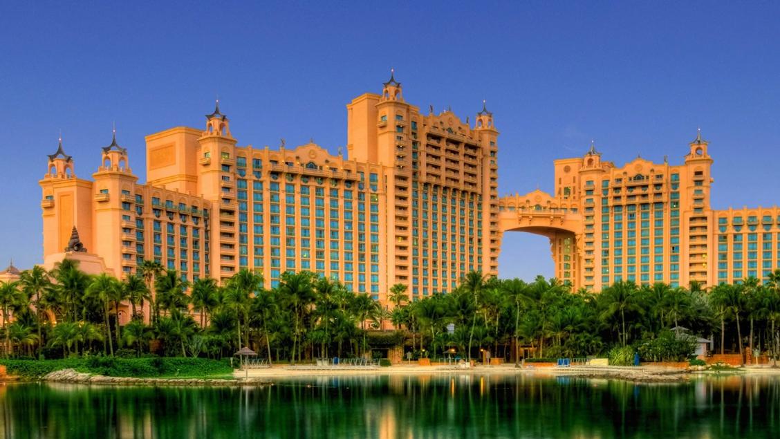 Travel X A large pink and blue resort building with multiple towers and a central archway, bordered by palm trees, reflects on a body of water in the foreground, creating an idyllic vacation setting. Discover your dream stay at Travel X's premier destination. Book Your Trip With Travel X