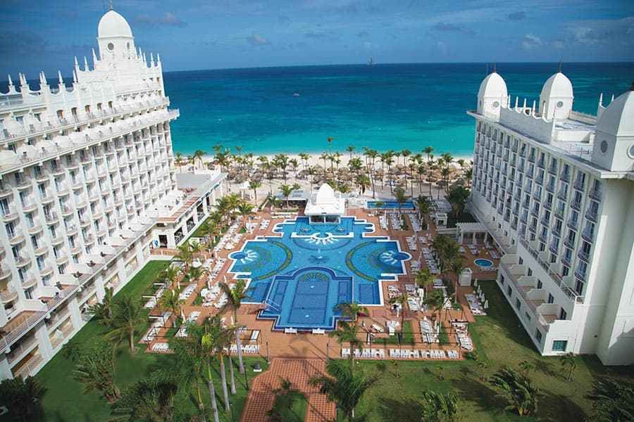 Travel X Aerial view of a luxury beachfront resort with two large white buildings, a central pool area, and a beach with clear blue waters in the background—perfect for your next vacation. Book your trip today! Book Your Trip With Travel X