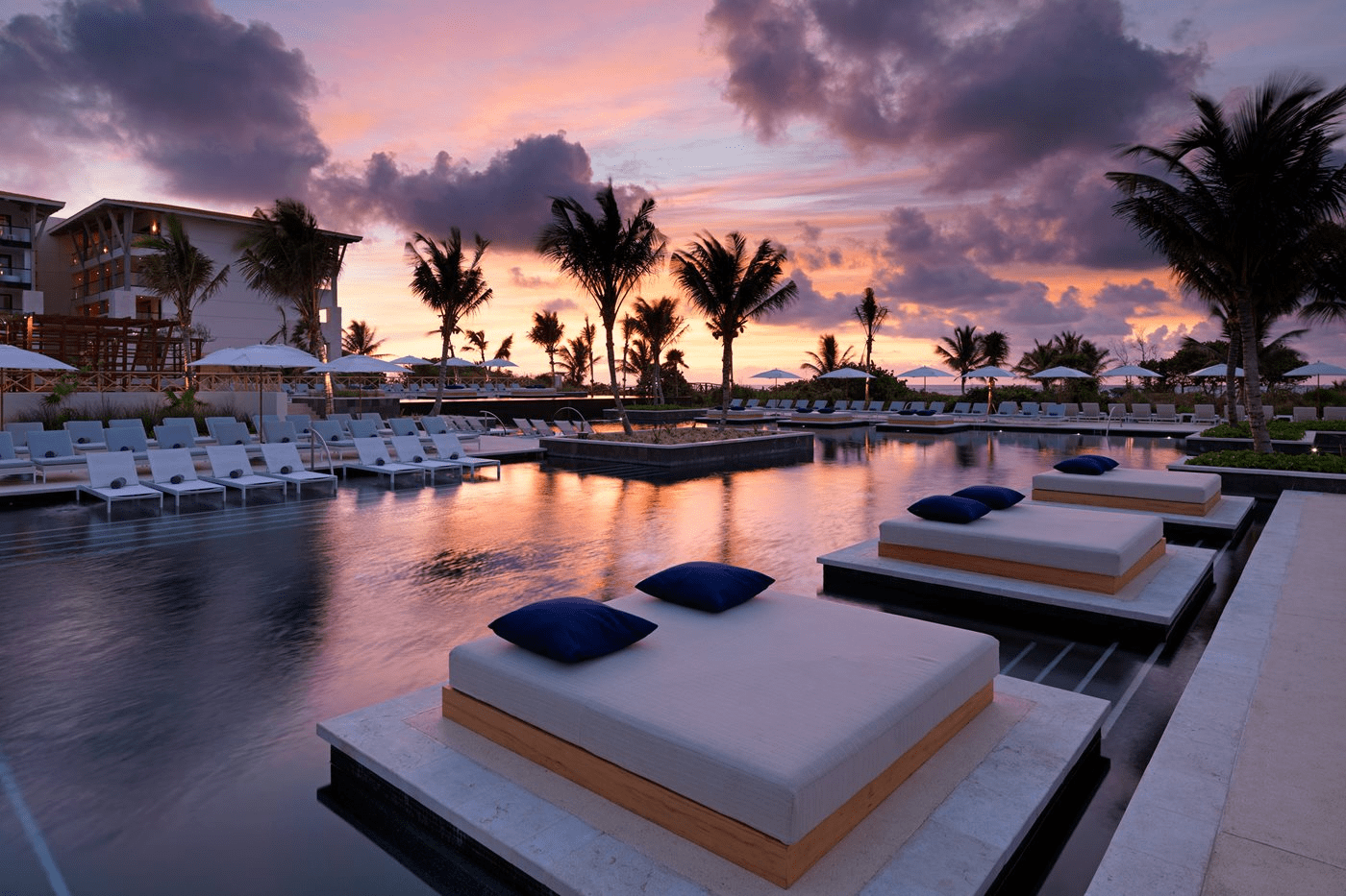 Travel X A luxurious outdoor pool area at sunset, featuring large cushioned lounge platforms with blue pillows, surrounded by palm trees and adjacent to a modern resort hotel. Perfect for your next vacation—book your trip now! Book Your Trip With Travel X