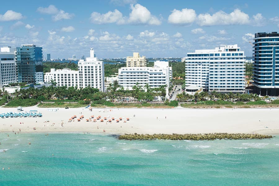 Travel X View of a coastline featuring sandy beaches, clear turquoise water, and several multi-story resort buildings and hotels under a partly cloudy sky. Beachgoers are seen scattered along the shore, enjoying their day. Book your trip with Travel X for an unforgettable beachside experience. Book Your Trip With Travel X