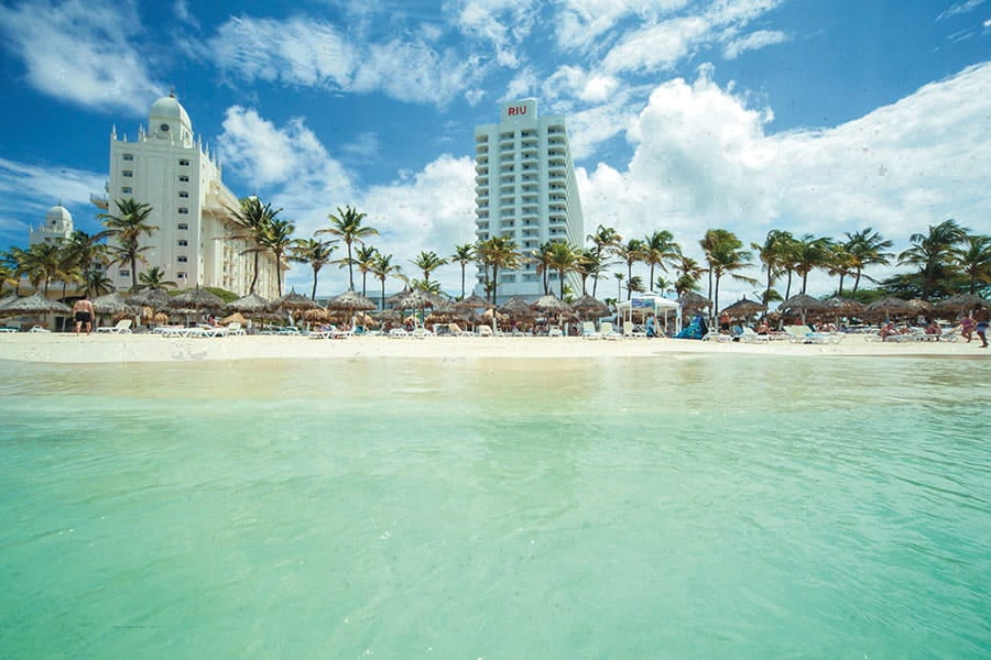 Travel X Beachfront view with two large resort hotels in the background, palm trees, beach huts, and people relaxing on the sand under clear, sunny skies. The ocean water is clear and calm. Experience paradise with Travel X—Book Your Trip today! Book Your Trip With Travel X