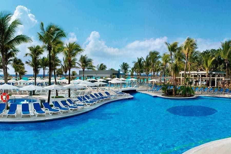 Travel X Experience the ultimate resort relaxation at our large outdoor pool area, complete with multiple lounge chairs, palm trees, and umbrellas under a clear blue sky. With the ocean visible in the background, it's time to book your trip with Travel X. Book Your Trip With Travel X