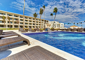 Travel X Relax by the outdoor pool area featuring wicker lounge chairs, a modern hotel building in the background, and tall palm trees under a partly cloudy sky. Experience this resort for yourself—Book Your Trip with Travel X today! Book Your Trip With Travel X