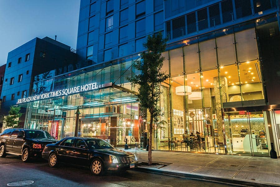 Travel X Nighttime view of the entrance to Riu Plaza New York Times Square Hotel, featuring glass walls and lit interiors with parked cars in front. Perfect for a resort-style vacation, book your trip now! Book Your Trip With Travel X