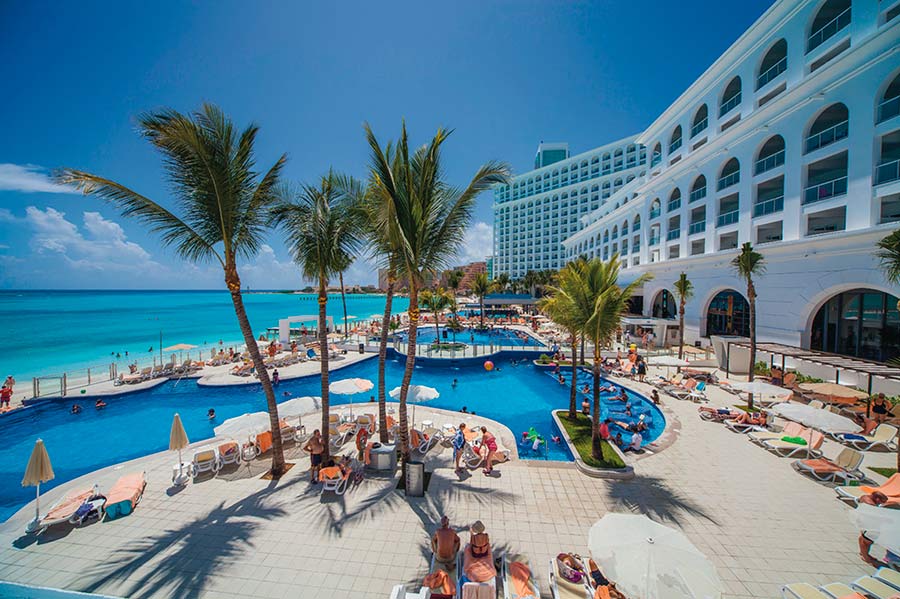 Travel X A resort with palm trees overlooks a large outdoor pool, surrounded by lounge chairs and umbrellas, adjacent to a turquoise ocean under a clear blue sky. Book your trip now for the ultimate vacation experience. Book Your Trip With Travel X