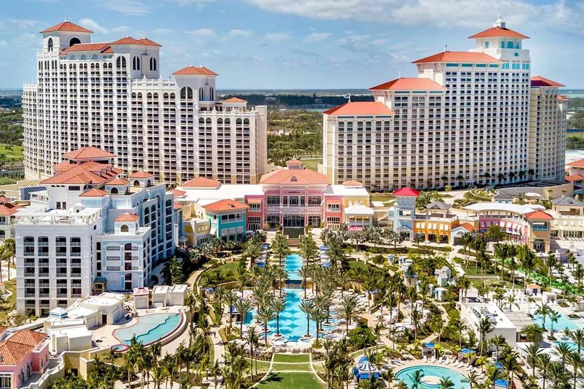 Travel X Aerial view of a large resort complex with multiple high-rise buildings, numerous pools, palm trees, and landscaped grounds under a partly cloudy sky. Book your trip now for an unforgettable vacation at this stunning resort. Book Your Trip With Travel X