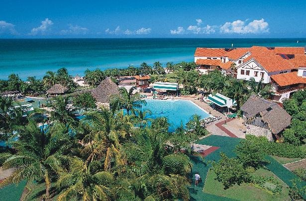 Travel X Aerial view of a seaside resort with a large swimming pool surrounded by palm trees, sun loungers, and buildings with red roofs. The ocean is visible in the background. Book your trip with Travel X for the perfect vacation experience! Book Your Trip With Travel X