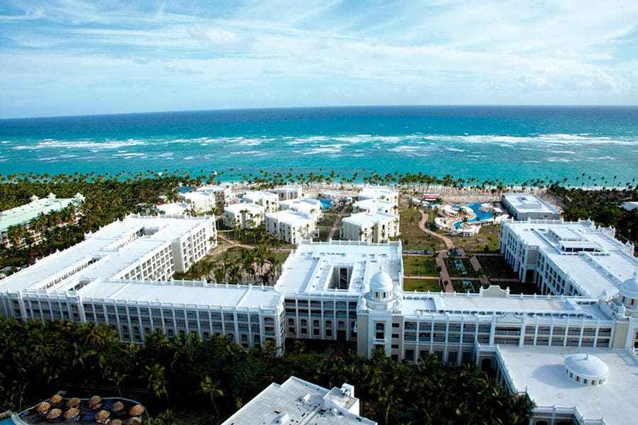 Travel X Aerial view of a large white beachfront resort complex with multiple buildings, situated next to the ocean with visible waves and surrounded by palm trees. Book your trip now with Travel X for an unforgettable stay. Book Your Trip With Travel X