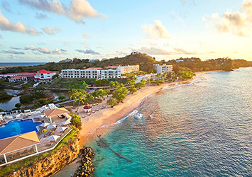 Travel X Aerial view of a beachfront resort with buildings, a swimming pool, sandy beach, and turquoise waters under a partly cloudy sky at sunset—book your trip for an unforgettable vacation. Book Your Trip With Travel X