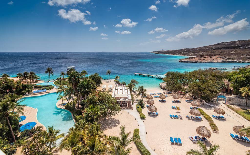 Travel X Aerial view of a tropical beach resort with multiple pools, palm trees, beach umbrellas, and a dock extending into clear blue ocean water. Hills and coastline are visible in the distance. Ready for your dream vacation? Book Your Trip now with Travel X! Book Your Trip With Travel X