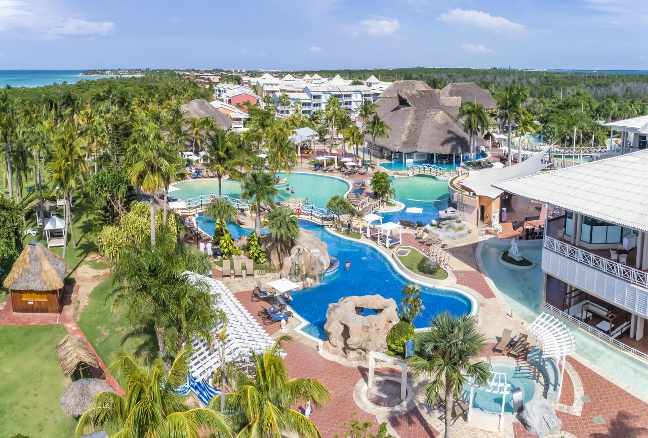 Travel X Aerial view of a tropical resort by Travel X with multiple swimming pools, palm trees, lounge chairs, and thatched-roof buildings, surrounded by lush greenery and adjacent to the coastline. Book your trip today for an unforgettable getaway! Book Your Trip With Travel X