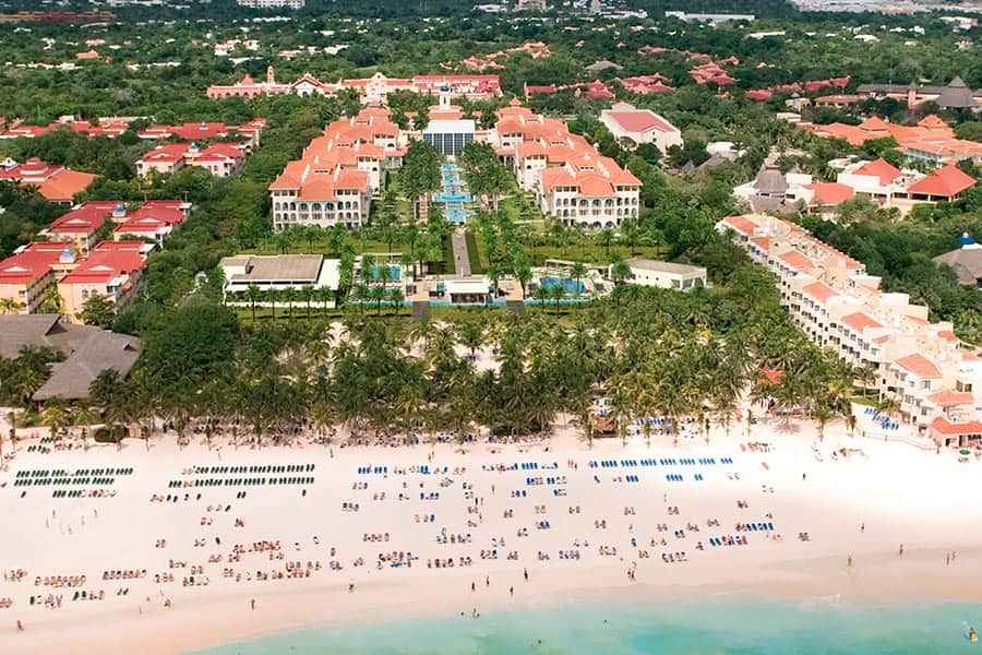 Travel X Aerial view of a beachfront resort with numerous loungers on a white sandy beach, surrounded by lush greenery and multiple buildings. Book your trip to this idyllic vacation spot today! Book Your Trip With Travel X