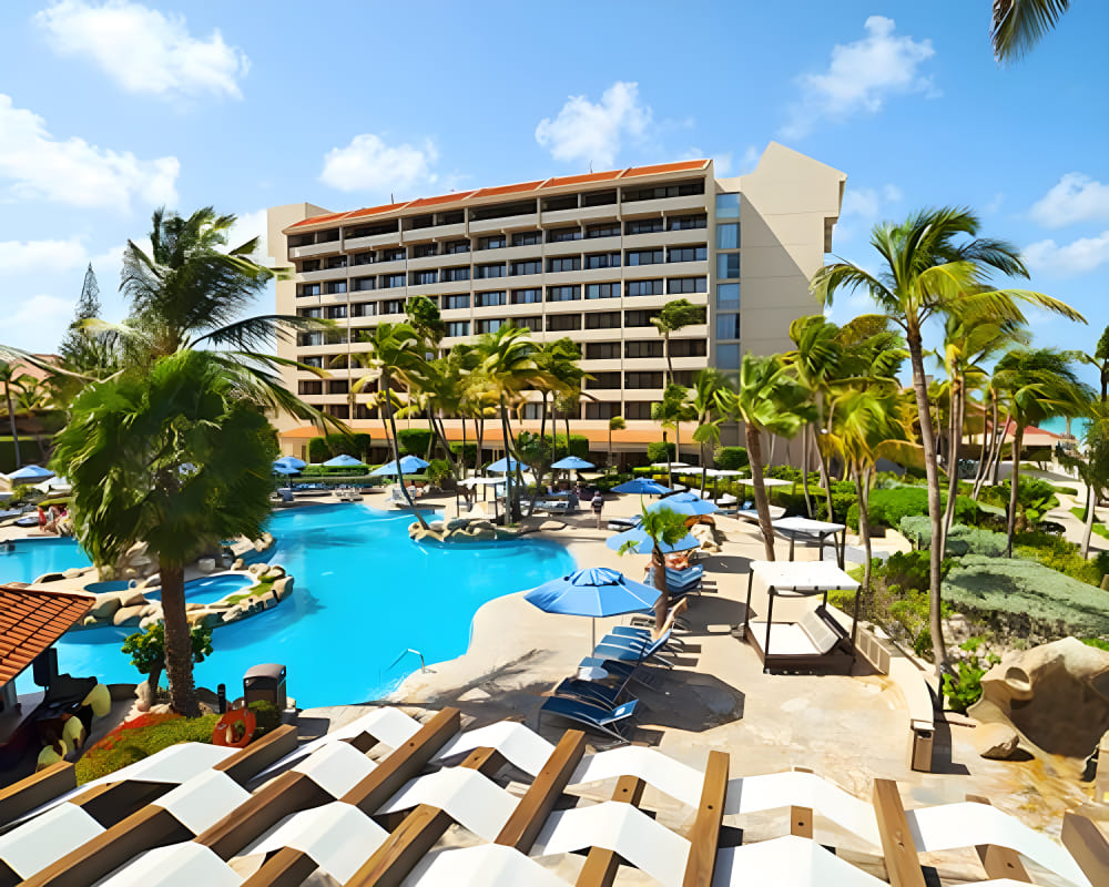 Travel X A vacation resort with a large outdoor pool surrounded by palm trees, lounge chairs, and sun umbrellas on a sunny day. Book your trip today! Book Your Trip With Travel X