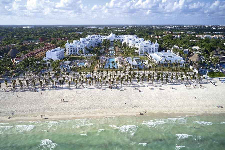 Travel X Aerial view of a beachfront resort with white buildings, multiple pools, palm trees, and loungers by the sea under a partly cloudy sky. Discover your dream vacation at Resort Travel X. Book your trip now! Book Your Trip With Travel X