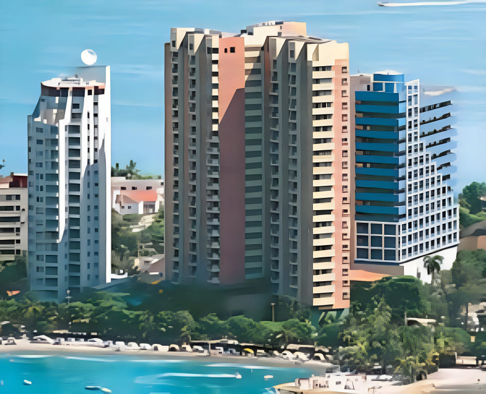 Travel X High-rise buildings stand along a beachfront, with smaller structures and greenery in the background. The foreground features a sandy beach with boats on the water, inviting you to book your trip today for an unforgettable stay at this coastal resort. Book Your Trip With Travel X