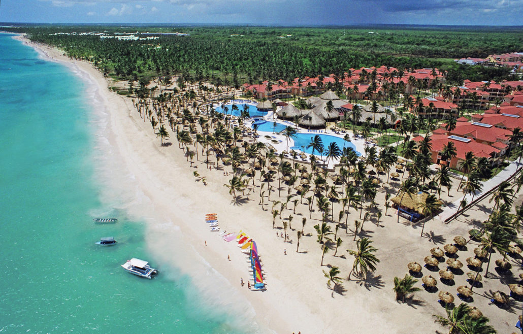 Travel X Aerial view of Travel X's tropical beachfront resort featuring turquoise water, sandy beach, numerous palm trees, several pools, and red-roofed buildings—a perfect vacation paradise. Book Your Trip With Travel X