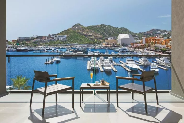 Travel X Two chairs and a table with a tea set on a balcony at the resort, overlooking a marina filled with boats and yachts, surrounded by mountains and buildings. Book Your Trip With Travel X