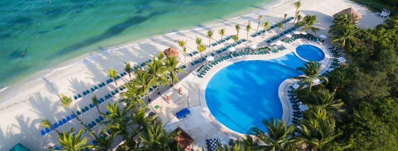 Travel X Aerial view of a beachfront resort with a large pool surrounded by lounge chairs and palm trees, adjacent to the blue-green ocean. Book Your Trip to this tropical paradise for an unforgettable vacation. Book Your Trip With Travel X