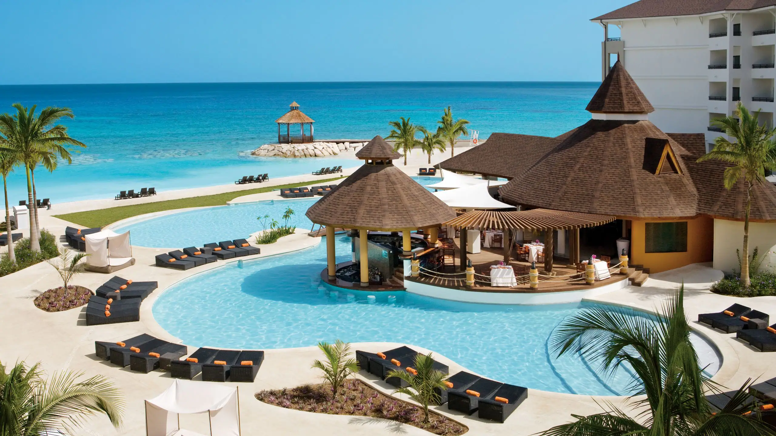 Travel X A tropical resort with a large pool, poolside cabanas, a swim-up bar, and a gazebo near the ocean under a clear blue sky. Book your trip with Travel X for the ultimate vacation. Book Your Trip With Travel X