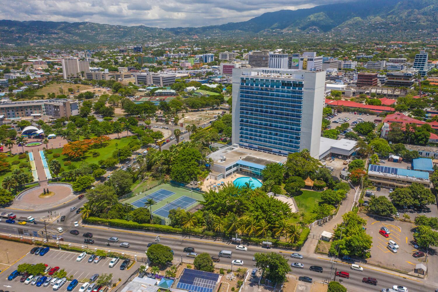 Travel X Aerial view of a cityscape with a tall modern building surrounded by trees, a swimming pool, and tennis courts. The area includes a busy road with cars and various buildings against a mountainous backdrop—a perfect scene to Book Your Trip with Travel X for an unforgettable resort experience. Book Your Trip With Travel X