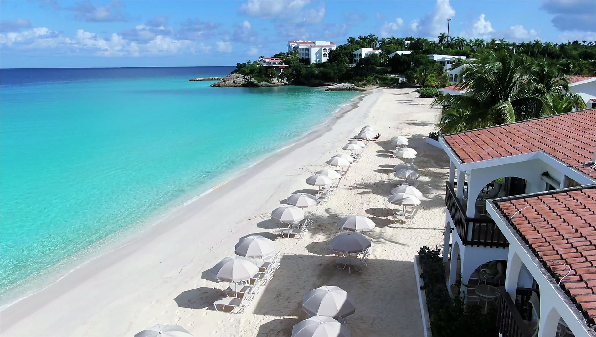 Travel X A sandy beachfront with rows of empty lounge chairs and umbrellas beside a turquoise sea, bordered by greenery and white buildings under a partly cloudy sky—an idyllic Travel X resort perfect for your next vacation. Book Your Trip With Travel X