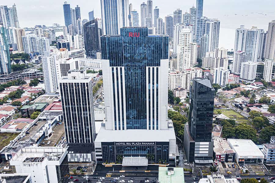 Travel X Aerial view of the Hotel Riu Plaza Panama, a tall modern building surrounded by various skyscrapers and city infrastructure in Panama City. Book your trip to this urban resort for an unforgettable vacation. Book Your Trip With Travel X