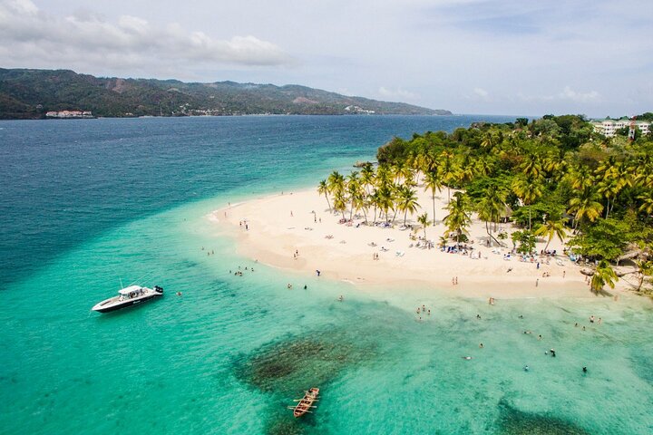 Travel X Aerial view of a tropical resort beach with light sand, turquoise water, palm trees, and people swimming and sunbathing. A boat is docked near the shore, and hills are visible in the background. Book your trip to this paradise for an unforgettable vacation experience! Book Your Trip With Travel X