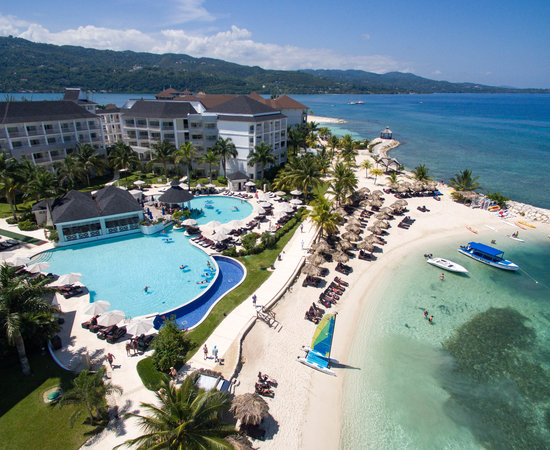 Travel X Aerial view of a Travel X beachfront resort featuring multiple pools, sun loungers, palm trees, and a docked boat in a stunning tropical setting. Perfect for your dream vacation. Book Your Trip With Travel X