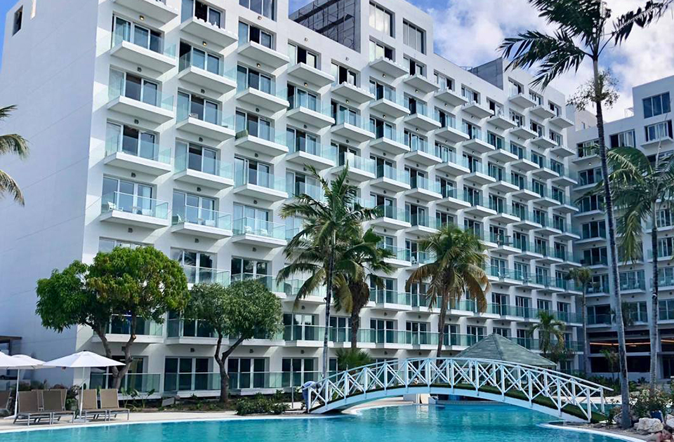 Travel X A large, multi-story resort with numerous balconies is situated next to a swimming pool. There are palm trees and a small footbridge over the pool, with lounge chairs and umbrellas nearby. Perfect for your next vacation—book your trip now! Book Your Trip With Travel X