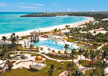 Travel X An aerial view of a tropical resort featuring a large swimming pool, surrounding palm trees, various buildings, and a sandy beach next to a turquoise ocean. Book your trip now for the ultimate vacation experience! Book Your Trip With Travel X