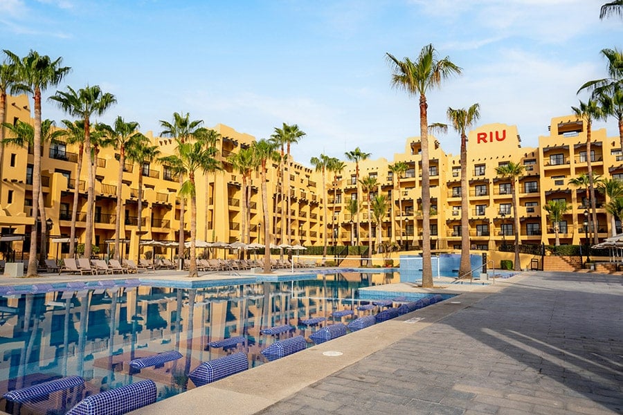 Travel X Outdoor poolside view of a RIU hotel with palm trees, lounge chairs, and a calm atmosphere. The hotel's multiple stories and beige facade are reflected in the clear pool water. Book your trip with Travel X for an unforgettable vacation experience. Book Your Trip With Travel X