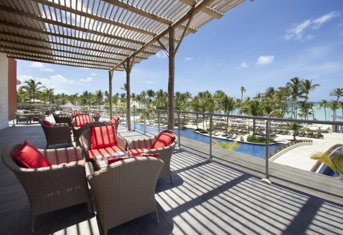 Travel X Outdoor seating area with wicker chairs and red cushions under a pergola, overlooking a pool and palm trees with a view of the ocean in the background. Perfect for a relaxing vacation, this spot embodies the charm of Travel X resorts. Book Your Trip With Travel X