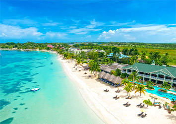 Travel X Aerial view of a tropical beach with clear turquoise water, white sand, beachfront resort buildings, palm trees, and sun loungers under umbrellas along the shore. Discover this paradise with Travel X and book your trip today! Book Your Trip With Travel X