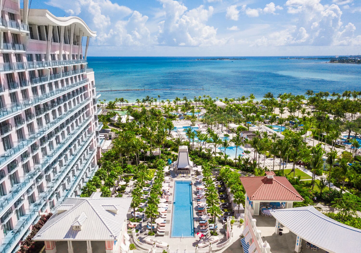 Travel X Aerial view of a beachfront resort with a large swimming pool, surrounded by palm trees and lounge chairs, adjacent to clear blue ocean waters and a bright, partly cloudy sky. Book your trip now for an unforgettable vacation experience! Book Your Trip With Travel X