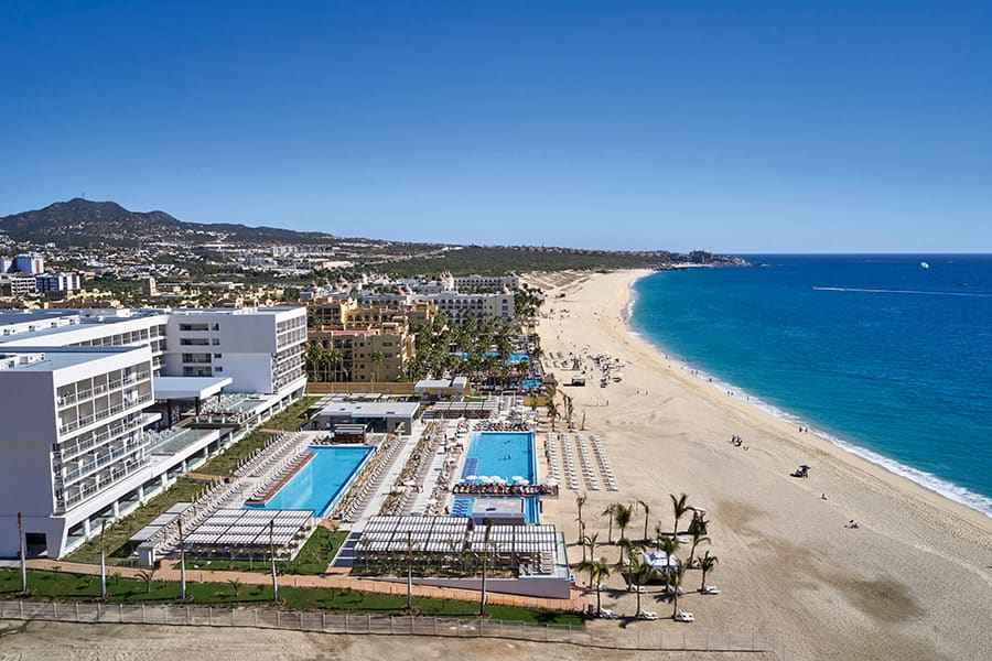 Travel X Aerial view of a coastal resort with several pools, lounge areas, and a sandy beach extending into the distance under a clear blue sky. Book your trip now for the ultimate vacation experience. Book Your Trip With Travel X