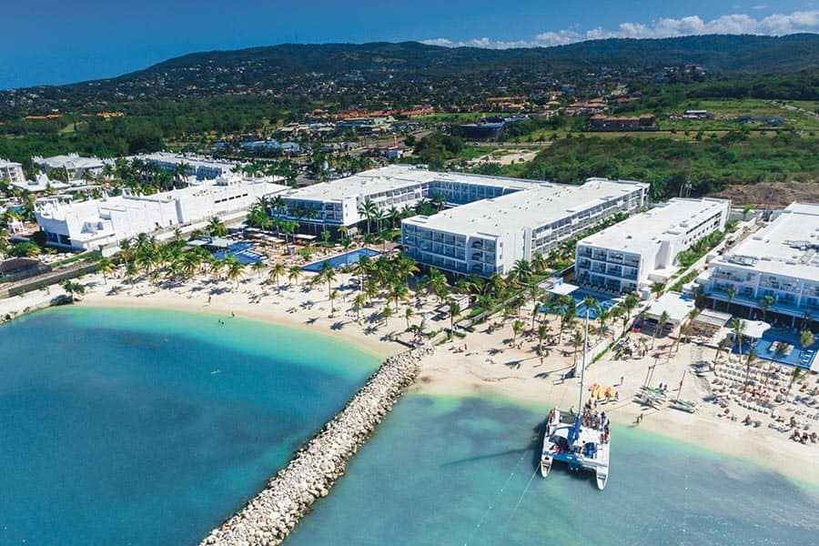 Travel X Aerial view of a coastal resort with multiple white buildings, palm trees, a sandy beach, a docked catamaran, and lush green hills in the background. Book your trip with Travel X for the ultimate vacation experience. Book Your Trip With Travel X